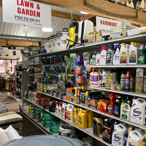 Store aisle with a variety of lawn and garden products on shelves, including fertilizers, weed killers, and tools. Signs above read 