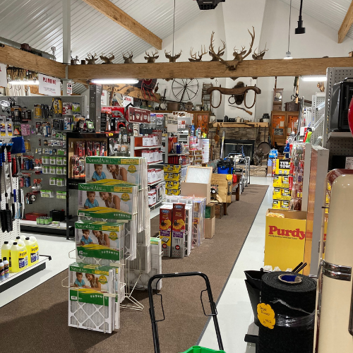 A cozy hardware store interior with shelves of tools, supplies, and paints. Antler mounts and wagon wheels decorate the high, rustic ceiling.