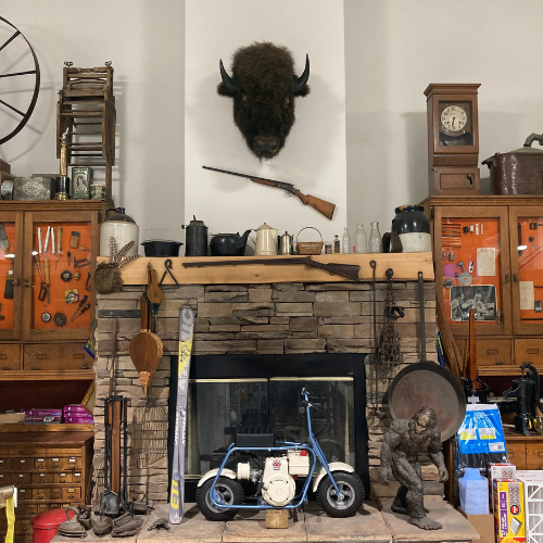 A rustic room features a stone fireplace with a bison head and rifle mounted above. Antiques and tools are displayed, creating a vintage, nostalgic ambiance.