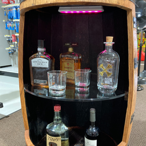 A wooden barrel shelf displays liquor bottles and glasses under pink LED lights. The setting is a retail store with tools visible in the background.