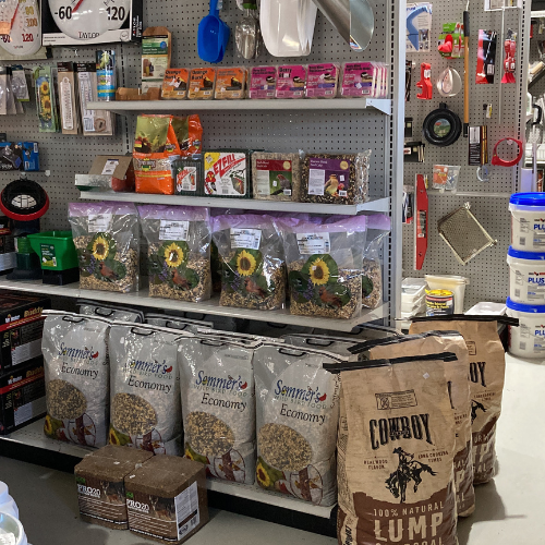 Hardware store aisle with shelves displaying gardening tools, birdseed bags, and charcoal. The scene conveys a busy, organized shopping space.