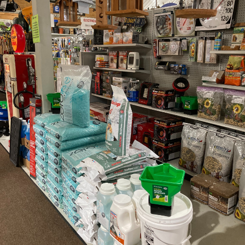 A hardware store aisle with various items for sale, including stacked bags of fertilizer, gardening tools, and shelves filled with bird seed and decor.