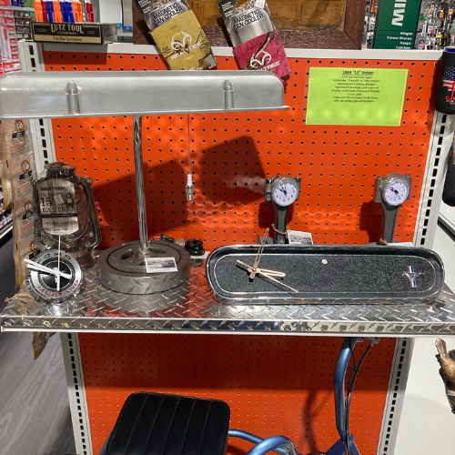 A display on a metallic shelf features a steel lamp, customized clock with vintage gauges, and industrial decor. A bright orange pegboard backdrop adds contrast.