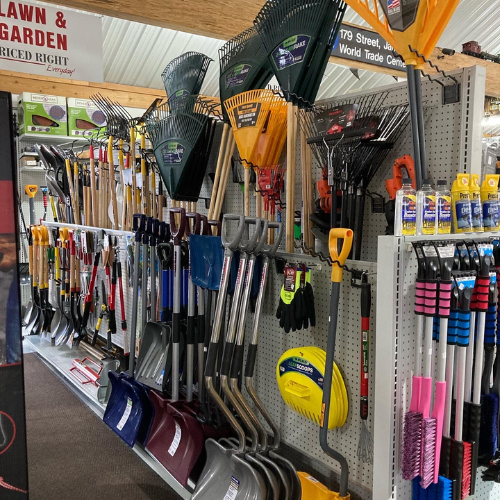 A hardware store aisle displays various gardening tools, including rakes, shovels, and brooms. Bright lighting emphasizes organization and utility.