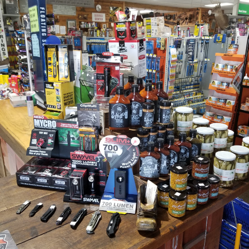 A wooden store counter displaying various items, including flashlights, sauces, and jars of pickles. Shelves with more products are visible in the background.