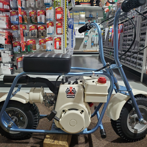 A blue and white mini bike is displayed in a hardware store. It features a small engine and a padded black seat. Shelves of tools and supplies are seen in the background.