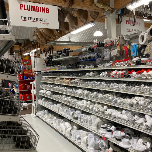Plumbing aisle in a hardware store with shelves filled with various white and gray fittings and pipes. Signs read 