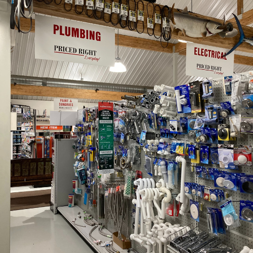 A hardware store aisle with plumbing and electrical supplies. Shelves are stocked with pipes, fittings, wires, and tools. Signs indicate 'Plumbing' and 'Electrical' sections, creating an organized and practical atmosphere.