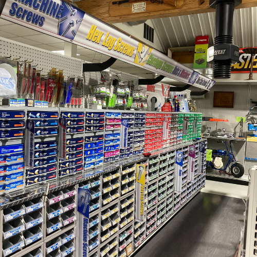 A well-organized hardware store aisle with shelves filled with boxes of screws in various colors. Signage overhead indicates machine and lag screws.