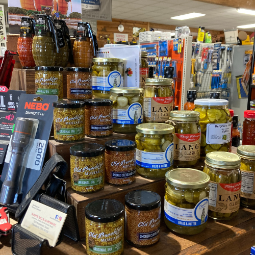 Rows of jars with pickles and mustard displayed on wooden shelves in a store, surrounded by flashlights and shop items, evoking a rustic market feel.