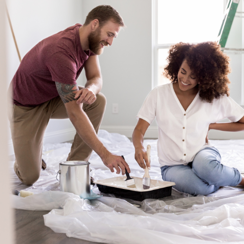 A couple kneels on a plastic-covered floor, smiling as they paint a room. A paint can and tray are beside them, with a ladder in the background.