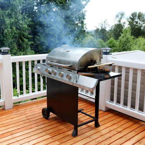 A silver grill on a wooden deck is steaming with a pan on the side, surrounded by lush green trees under a partly cloudy sky, evoking tranquility.