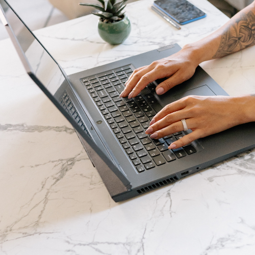 Hands typing on a laptop keyboard atop a white marble table, next to a small potted plant and a smartphone. The scene conveys focus and modernity.