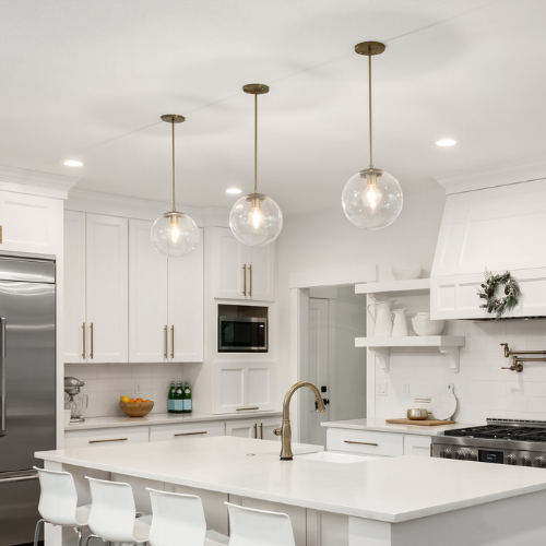 Modern kitchen with a white island and bar stools, featuring pendant lights, stainless steel appliances, and a touch of greenery above the stove.