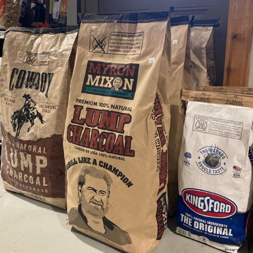 Bags of charcoal varieties: Cowboy and Myron Mixon’s lump charcoal with rustic designs, and Kingsford briquettes with a classic logo, lined up indoors.