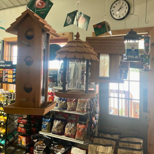 Hanging wooden bird feeders in a rustic store setting with bags of pet food on shelves. A clock and gardening items are visible in the background.