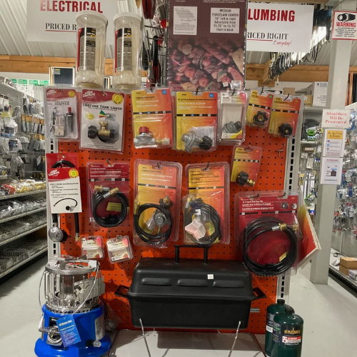A store display features a variety of camping accessories on an orange pegboard. Items include propane tank gauges, hoses, and a lantern heater.