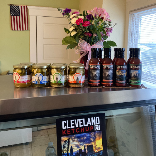 Jars of pickles and bottles of Cleveland Ketchup on a countertop, with a bouquet of flowers in the background and an American flag on the wall.