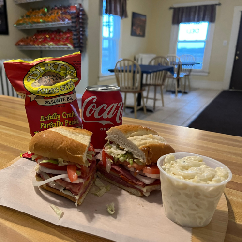 A deli sandwich with layers of meats, lettuce, and tomato sits on a table next to potato salad, a Coca-Cola can, and a snack bag. Bright, casual dining room.