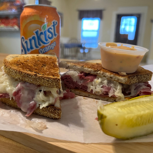 A Reuben sandwich with corned beef, sauerkraut, and cheese on rye is cut in half. It’s paired with a pickle slice, a dipping sauce, and a can of Sunkist Orange.