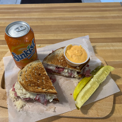 Grilled sandwich with corned beef and sauerkraut, a small cup of orange sauce, dill pickles, and a can of Sunkist on a wooden table.