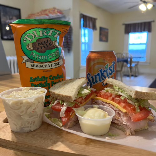 A wooden tray holds a turkey sandwich with lettuce and tomato, a cup of potato salad, Sriracha Honey chips, and a can of Sunkist, set in a cozy room.