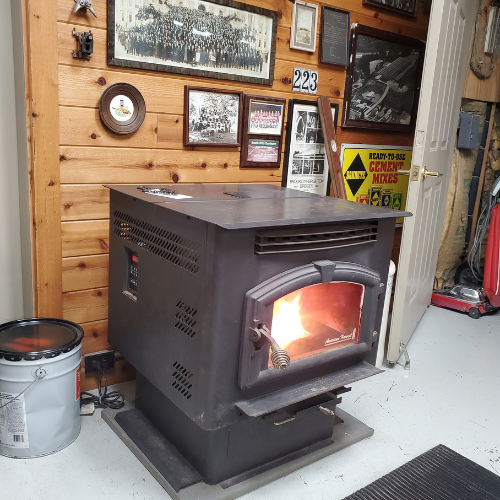 A cozy wooden room features a glowing black stove emitting warmth. Vintage photographs and a colorful poster adorn the walls, creating a nostalgic ambiance.