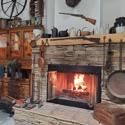 A cozy rustic room with a stone fireplace ablaze, surrounded by antique items including a rifle, teapots, and wooden furniture, creating a warm vintage ambiance.