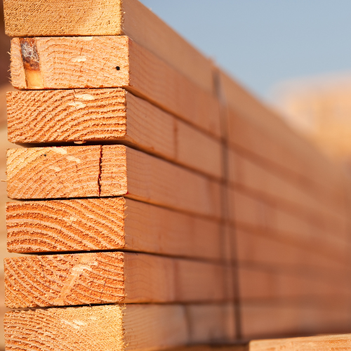 Stack of neatly arranged, light brown wooden planks lined vertically, showing natural wood grain and textures, under soft, warm daylight.